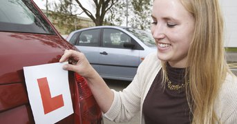 Female leaner driver handles L-plate at rear of car, day.