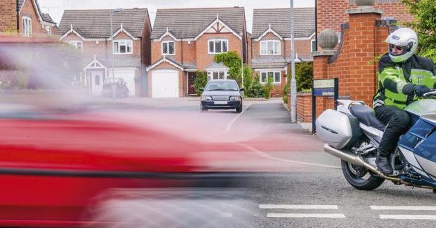Car passes junction in a blur as motorcylce rider pauses, waiting to emerge onto main road.