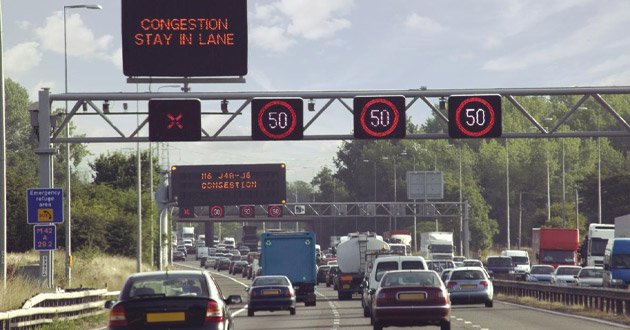Motorway congestion speed limiter sign illuminated over heavy traffic, day.